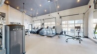 This modern fitness center is a thoughtfully designed exercise space that offers a comprehensive workout environment. The room is defined by its striking high ceilings, finished in a bold dark gray, which create a dramatic contrast against the bright white walls and light-toned flooring. Exposed industrial-style ductwork and recessed lighting add to the contemporary aesthetic throughout the space.

Large, glass-panel garage-style doors line one wall, flooding the interior with abundant natural light and providing open views of the surrounding area, contributing to an energetic and airy atmosphere. A motivational "Hustle for the Muscle" wall display serves as a bold focal point, reinforcing the space's active purpose.

The fitness center is fully equipped with a wide range of True-brand cardio machines, including treadmills, ellipticals, stair climbers, rowing machines, and stationary bikes. A dedicated free weights area features a full dumbbell rack, while multiple strength and cable machines are arranged throughout the open floor plan, allowing for efficient use of the space.

Large wall mirrors expand the sense of space and support proper form during workouts. Floor-to-ceiling glass doors connect the fitness center to an adjacent lounge or lobby area, enhancing the flow between spaces. The overall design balances industrial character with functionality, creating an inviting and well-appointed fitness destination.