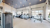 This modern fitness center is a thoughtfully designed exercise space that offers a comprehensive workout environment. The room is defined by its striking high ceilings, finished in a bold dark gray, which create a dramatic contrast against the bright white walls and light-toned flooring. Exposed industrial-style ductwork and recessed lighting add to the contemporary aesthetic throughout the space.

Large, glass-panel garage-style doors line one wall, flooding the interior with abundant natural light and providing open views of the surrounding area, contributing to an energetic and airy atmosphere. A motivational "Hustle for the Muscle" wall display serves as a bold focal point, reinforcing the space's active purpose.

The fitness center is fully equipped with a wide range of True-brand cardio machines, including treadmills, ellipticals, stair climbers, rowing machines, and stationary bikes. A dedicated free weights area features a full dumbbell rack, while multiple strength and cable machines are arranged throughout the open floor plan, allowing for efficient use of the space.

Large wall mirrors expand the sense of space and support proper form during workouts. Floor-to-ceiling glass doors connect the fitness center to an adjacent lounge or lobby area, enhancing the flow between spaces. The overall design balances industrial character with functionality, creating an inviting and well-appointed fitness destination.