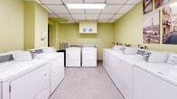 Located at 3777 Rue Saint-Urbain in Montréal's vibrant Mile End neighbourhood, this shared laundry facility is a practical and neatly organized amenity space. The room features a dropped acoustic tile ceiling with integrated fluorescent lighting that provides bright, even illumination throughout. The walls are painted in a light yellow-green tone, lending the space a clean and welcoming atmosphere.

Multiple top-loading washing machines and front-loading dryers are arranged along the perimeter walls, offering ample capacity for residents. The machines are coin-operated and appear well-maintained, providing reliable laundry services. A hand sanitizer dispenser is mounted near the entrance, and instructional signage is posted to guide users through proper machine operation.

The flooring consists of light-toned tile that is easy to clean and contributes to the room's overall brightness. Decorative framed prints featuring Parisian street scenes and architectural imagery are displayed on the walls, adding a thoughtful aesthetic touch to an otherwise functional space. A framed mirror also adorns one wall, further enhancing the room's visual appeal. Access is provided through a solid door, ensuring a secure and private environment. The space is efficiently laid out to maximize usability while maintaining clear pathways between machines.