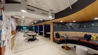 This contemporary single-floor office space is designed to accommodate a wide range of work styles through a thoughtfully zoned layout. The floor plan reveals a generous open area anchored by collaborative workstations, semi-private booth seating, and enclosed glass-walled meeting rooms that promote transparency while maintaining acoustic separation.

A standout feature is the curved lounge alcove, finished with a warm wood ceiling, deep navy walls adorned with a lush living plant wall of succulents and air plants, and plush wraparound banquette seating in tan velvet. Black marble-topped tables on brass pedestal bases lend the space an elevated, hospitality-inspired aesthetic.

Throughout the open floor, modular work tables on geometric patterned rugs provide flexible group seating, while navy-blue private work booths offer quieter individual focus zones. A bold, colorful geometric accent wall adds artistic energy to the corridor.

Additional amenities include a beverage station with under-counter refrigeration, lounge sofas dressed in teal and graphic-print cushions, and glass-enclosed offices equipped for meetings or individual work. The exposed ceiling infrastructure is painted in crisp white, complementing the dark charcoal accent walls and maintaining an industrial-modern character. Recessed and track lighting throughout ensures a well-lit, professional environment.