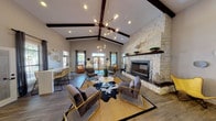 Located at 3903 SE Military Drive in San Antonio, Texas, this single-story community clubhouse and leasing office combines modern Texas Hill Country aesthetics with contemporary design elements. The main lounge area immediately draws attention with its soaring vaulted ceiling accented by dark wood exposed beams and an eye-catching multi-globe brass chandelier that serves as a central focal point. A floor-to-ceiling natural limestone fireplace with a mosaic tile surround and a dark wood mantel anchors the space, creating a warm and inviting atmosphere. The seating area is thoughtfully arranged with a mix of velvet accent chairs, leather loungers, and a cowhide rug layered over woven textiles, reflecting an eclectic, curated style. Large windows dressed with sheer curtains and blinds allow abundant natural light to fill the interior while offering views of the surrounding outdoor courtyard. Wood-look plank flooring runs throughout, providing a cohesive and durable foundation. A dedicated leasing office area features dark-stained desks, modern chrome-framed chairs, and bright windows that maintain an open, professional atmosphere. An additional media or multipurpose room showcases a built-in white cabinetry wall unit with an integrated flat-screen display and vaulted ceiling with exposed beams, completing this well-appointed community facility.