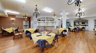 Located at 3980 S Jackson Dr in Independence, Missouri, this senior living facility offers a generously sized, single-floor layout with a welcoming and elegantly appointed interior. The grand entrance lobby immediately impresses with its soaring two-story ceiling, an upper-level balcony adorned with lush greenery, and a staffed reception desk, setting a warm and professional tone for the entire facility. Patterned, neutral-toned carpeting flows throughout the common areas and hallways, providing both comfort and visual cohesion. The spacious hallways are thoughtfully decorated with wall art, ornamental sconces, wall-mounted displays, and decorative niches featuring floral arrangements, creating an inviting residential atmosphere. A comfortable lounge area features leather seating, a chandelier, column accents, and built-in shelving, offering residents a refined space to relax. The dining area is furnished with multiple tables and chairs beneath warm lighting, ideal for communal gatherings. Additional gathering spaces with columned archways and skylight features provide bright, open areas for socialization. A designated area marked "Wine Cellar" adds a distinctive character to the facility. Throughout, rich wood-toned doors and chair rails complement the neutral wall tones, contributing to the overall warm and dignified ambiance of this well-appointed space.