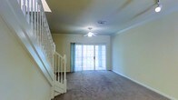 This two-story home offers a thoughtfully designed layout that balances comfort and functionality. The main living area is anchored by plush wall-to-wall carpeting that adds warmth underfoot, while crown molding along the ceiling lends a refined architectural touch. A ceiling fan with a built-in light fixture provides both ventilation and ambient lighting, making the space practical year-round. Large sliding glass doors dominate one wall of the living area, allowing natural light to pour in and offering direct access to the outdoor area beyond — a standout feature that enhances the sense of openness and connection to the exterior. Sheer vertical blinds frame the sliding doors, offering adjustable light control and privacy. A classic white staircase with decorative spindles and a sturdy balustrade rises gracefully from the main level, serving as both a functional and visually appealing transition to the upper floor. The neutral color palette throughout — from the soft beige walls to the muted carpet tones — creates a calm, versatile backdrop well-suited to a variety of interior styles. This home's two-floor design provides ample separation between living and private spaces, making it a well-rounded and inviting residence.