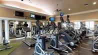 This fitness center offers a thoughtfully designed workout space accommodating a robust selection of cardio equipment. Rows of treadmills and elliptical machines, including Star Trac models, are arranged to maximize floor space while maintaining an open, accessible feel. Each machine is equipped with its own integrated display screen, and additional wall-mounted flat-screen televisions are positioned at eye level along the upper walls, ensuring entertainment options during workouts. The interior features warm, neutral-toned walls accented by white crown molding, lending the space a polished, upscale aesthetic. A ceiling fan provides additional air circulation overhead, complementing the recessed lighting that evenly illuminates the room. Full-length mirrors line portions of the walls, visually expanding the space and allowing users to monitor their form. Decorative columns add architectural character while subtly defining different zones within the room. Natural light filters in through windows on one side, contributing to a bright and energizing atmosphere. The dark rubberized flooring provides both durability and safety underfoot.