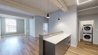 Located at 160 Lafayette Street in Jersey City, NJ, this one-bedroom, one-bathroom unit showcases a compelling blend of industrial character and modern finishes. The open-concept kitchen and living area serves as the heart of the home, anchored by a two-tone peninsula with a crisp white quartz countertop and slate-gray cabinetry. The kitchen is well-appointed with stainless steel appliances, including a refrigerator, range, and microwave, complemented by a mosaic tile backsplash and upper white cabinetry. Pendant lighting and exposed ceiling beams and piping reinforce the loft-inspired aesthetic throughout the living area. Light wood-plank flooring flows seamlessly across the entire unit, enhancing the sense of openness and warmth. The spacious bedroom features painted exposed brick walls, original arched windows that invite natural light, and overhead structural beams that add architectural depth. The bathroom is finished with clean white tile surrounds on the tub/shower combo, gray mosaic accent tile, a white quartz vanity top, and slate-gray cabinetry consistent with the unit's design language. A dedicated laundry area and closet round out the practical amenities, making this an efficient and stylish urban residence.