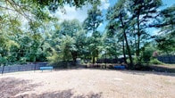 This outdoor space is a dedicated dog park enclosed by black metal fencing, providing a secure and open area for pets to roam freely. The ground is covered with natural dirt and pine straw, creating a low-maintenance surface well-suited for active use. Two blue benches are positioned along the perimeter, offering comfortable seating for pet owners while they supervise their animals.

The park is framed by a dense canopy of mature pine and deciduous trees, which provide generous natural shade and lend the space a tranquil, wooded atmosphere. The tree cover helps moderate temperatures and creates a pleasant environment during warmer months. The lush greenery surrounding the fenced boundary adds a sense of privacy and seclusion, making the area feel like a peaceful retreat within a natural setting.

The fencing runs the full perimeter of the space, ensuring pets remain safely contained. An informational sign is posted near the center of the fence line, likely outlining park rules or guidelines. Overall, the space is thoughtfully designed with both pet safety and owner comfort in mind, set against a naturally scenic backdrop.