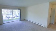 This single-floor space features a simply designed living area finished with neutral wall tones and wall-to-wall carpeting that lends a clean, understated aesthetic. The room is generously proportioned, providing a comfortable sense of openness. A large sliding glass door serves as a prominent architectural feature, allowing abundant natural light to fill the interior while offering direct views of a lush, green outdoor area. The sliding door is dressed with sheer curtains that soften incoming light while maintaining a connection to the outdoors. Along the base of one wall, a continuous baseboard heating unit runs the length of the room, providing an efficient and unobtrusive heating solution. The smooth, light-colored walls remain largely unadorned, offering flexibility for personalization. The overall ambiance is calm and neutral, making the space highly adaptable for a variety of furnishing styles and functional uses. The combination of natural light, outdoor access, and practical heating infrastructure makes this a functional and inviting living environment.