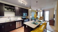 This 1-bedroom, 1-bathroom residence offers a thoughtfully designed single-level layout that maximizes both function and style. The kitchen is a standout feature, showcasing rich dark espresso cabinetry, granite countertops, a mosaic tile backsplash, and a full suite of stainless steel appliances including a refrigerator, electric range, microwave, and dishwasher. A granite-topped island with pendant lighting provides additional prep space and doubles as a casual dining area with seating. Wood-style flooring runs throughout the kitchen and entry, adding warmth and durability.

The open layout connects the kitchen seamlessly to the living area, which is anchored by a bold mustard-yellow accent wall and a ceiling fan. Natural light filters through windows dressed with patterned drapes and blinds, creating a bright and inviting atmosphere.

The bedroom is a serene retreat featuring a striking teal accent wall, a ceiling fan, carpeted flooring, and windows that offer natural light and outdoor views. A door leads directly into the bathroom, which features dark cabinetry, a granite vanity, a large mirror with vanity lighting, and wood-style flooring. The bathroom also provides access to an in-unit laundry area with washer and dryer connections visible in the mirror's reflection.