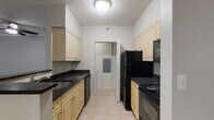 Located at 325 Speen St in Natick, Massachusetts, this single-floor residential unit offers a well-organized layout with a variety of functional living spaces. The galley-style kitchen is equipped with light wood cabinetry, dark granite countertops, black appliances including a refrigerator, electric range, built-in microwave, and dishwasher, all set atop neutral tile flooring that runs the length of the kitchen corridor. The living area features rich, dark hardwood-style flooring, a ceiling fan with light fixture, crown molding, and large windows with vertical blinds that allow natural light to fill the room. The carpeted bedrooms offer a comfortable feel, with one room featuring recessed lighting and a window with exterior views, and another providing access to a walk-in closet and an adjoining full bathroom. The bathrooms are finished with a classic black-and-white mosaic tile floor, white subway tile surrounding the soaking tub and shower combination, a dark granite vanity top with an undermount sink, white cabinetry, and a large mirror with overhead lighting. Additional bedroom spaces feature plush carpeting, recessed lighting, and windows that bring in natural light, rounding out a practical and cohesive living environment.