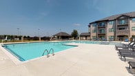 Located at 4000 SW Modern Way in Bentonville, Arkansas, this modern residential community blends contemporary architecture with resort-style amenities. The exterior showcases a multi-story building finished with a combination of warm siding and stone accents, complemented by private balconies that overlook the well-appointed outdoor spaces.

The centerpiece of the outdoor area is a generously sized swimming pool with crystal-clear blue water, surrounded by a broad concrete sun deck lined with sleek chaise lounges. A separate wading area sits adjacent to the main pool, and a volleyball net adds a recreational touch. A covered gazebo structure nearby provides shaded gathering space, while a dedicated lounge seating area features a large cantilever umbrella and curved sectional sofa — ideal for outdoor relaxation.

Inside, the grand lobby makes a striking first impression with soaring ceilings, large-format stone-look tile flooring, and a prominent stone pillar housing a modern fireplace. Lush tropical planters and curated seating arrangements contribute to an inviting, upscale atmosphere. Glass railings above hint at the open, airy vertical design of the building's interior. A bar-height seating area with stylish upholstered stools offers a casual spot for residents to gather. Throughout the community, the design emphasizes clean lines, natural materials, and an elevated sense of contemporary living.