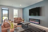 This single-floor space is thoughtfully designed to serve as a multifunctional gathering area, combining entertainment, dining, and relaxation zones in one open, cohesive layout. The interior features light hardwood-style flooring throughout, recessed lighting, and a neutral color palette accented by bold slate-blue feature walls that add depth and character.

The game room is a focal point, centered around a substantial pool table beneath a trio of industrial pendant lights. Adjacent to it, a comfortable lounge area features rich cognac leather sofas arranged around a sleek wall-mounted television and a modern linear electric fireplace, providing a warm, inviting atmosphere. Windows allow natural light to filter into the space.

The kitchen is well-appointed with dark wood cabinetry, granite countertops, stainless steel appliances including a refrigerator and dishwasher, and a large central island with bar seating. Additional counter-height seating lines one wall, offering flexible dining options.

A separate dining nook includes a rustic reclaimed wood table and benches, accented by wall-mounted planters and eclectic license plate wall art, adding a playful, informal touch to the overall design.