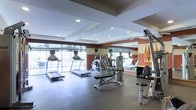 This fitness center offers a thoughtfully designed workout environment spread across a single floor. The space is anchored by a row of cardio machines, including treadmills and an elliptical trainer, positioned directly in front of expansive floor-to-ceiling windows that flood the room with natural light and provide views of the surrounding outdoor environment. The open layout ensures ample space between equipment, allowing for comfortable movement throughout the facility. A full-length mirrored wall runs along one side of the room, enhancing the sense of spaciousness while allowing users to monitor their form during workouts. The strength training area includes a multi-function cable and weight machine along with additional resistance equipment, catering to a range of exercise routines. Recessed lighting in the coffered ceiling supplements the natural light, ensuring the space remains bright and well-lit at all times. The flooring is finished in durable, dark rubber matting, practical for high-traffic fitness use. The walls feature a warm color palette of deep red and neutral tones, giving the space a modern and energizing aesthetic. Overall, this fitness center combines functionality with a clean, contemporary design.