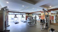 This fitness center offers a thoughtfully designed workout environment spread across a single floor. The space is anchored by a row of cardio machines, including treadmills and an elliptical trainer, positioned directly in front of expansive floor-to-ceiling windows that flood the room with natural light and provide views of the surrounding outdoor environment. The open layout ensures ample space between equipment, allowing for comfortable movement throughout the facility. A full-length mirrored wall runs along one side of the room, enhancing the sense of spaciousness while allowing users to monitor their form during workouts. The strength training area includes a multi-function cable and weight machine along with additional resistance equipment, catering to a range of exercise routines. Recessed lighting in the coffered ceiling supplements the natural light, ensuring the space remains bright and well-lit at all times. The flooring is finished in durable, dark rubber matting, practical for high-traffic fitness use. The walls feature a warm color palette of deep red and neutral tones, giving the space a modern and energizing aesthetic. Overall, this fitness center combines functionality with a clean, contemporary design.