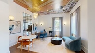 Located at 1200 River Dr in Belmont, NC, this thoughtfully designed single-floor space offers a generous and well-appointed layout comprising a living room and connecting hallway. The living room is the centerpiece of the space, showcasing rich hardwood flooring, elevated ceilings with a distinctive teal-toned textured finish, and exposed wooden beams that lend a warm, rustic character. A structural brick column adds an architectural focal point, complementing the eclectic interior design aesthetic throughout. Multiple large windows dressed with sheer gray curtains flood the interior with natural light while framing views of the surrounding outdoors. The living area is thoughtfully organized to accommodate both lounge and dining functions, with a built-in cabinetry wall featuring glass-front display cases and a mirrored backdrop serving as an elegant design anchor. Recessed lighting and statement light fixtures, including a gold flush-mount ceiling piece, enhance the ambiance. A secondary room off the hallway features a vibrant painted ceiling, wall sconces, carpeted flooring, and its own window view — ideal as a flex or office space. The hallway connects the areas seamlessly, providing a smooth flow throughout. Overall, the space exudes a curated, upscale character with a blend of classic and contemporary design elements.