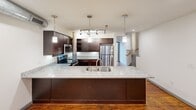This two-story loft apartment in Charlotte's vibrant Plaza Midwood neighborhood showcases an impressive mix of industrial charm and contemporary upgrades. The main level features rich hardwood floors, exposed brick accent walls, and structural columns that speak to the building's historic character. The open kitchen is a standout, boasting granite countertops, a large island with a double sink, dark espresso cabinetry, stainless steel appliances, and stylish pendant lighting. The bathroom includes granite counters, a rustic wood beam detail, and a clean tub-shower combo. The upper level provides a more intimate space, accessible via a staircase visible from the main living area. Large windows allow natural light to pour in, highlighting the warm hardwood floors and exposed brick throughout.