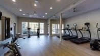 This single-floor fitness center offers a bright, welcoming environment designed for effective workouts. The space is finished with smooth hardwood-style flooring that runs throughout, providing a clean and polished aesthetic. Large windows line the walls, flooding the interior with natural light and offering pleasant views of the surrounding greenery, creating an energizing atmosphere for exercise.

The layout is open and spacious, allowing for comfortable movement between equipment. A row of modern treadmills with individual display screens is positioned along one side of the room, while stationary bikes and additional cardio equipment occupy the remaining floor space. Structural columns are integrated into the design, subtly defining different zones within the fitness area without disrupting the overall flow.

Ceiling fans are distributed throughout the space to maintain comfortable air circulation during workouts, complemented by recessed lighting that ensures even illumination regardless of the time of day. Neutral-toned walls contribute to the clean, contemporary feel of the room. Overall, the fitness center balances functionality and comfort, making it a practical and inviting space for daily exercise.