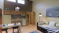This single-floor studio is defined by its striking architectural proportions, most notably the dramatic double-height ceilings that create an expansive, airy atmosphere. Clerestory windows positioned high on the walls draw in generous natural light, brightening the entire space throughout the day.

The kitchen is efficiently designed with light wood cabinetry, a white gas range, a double sink, and ample counter space. A pendant light hangs above the adjacent dining area, which features a compact table with bar-style seating — an ideal setup for casual meals. The kitchen flows seamlessly into the living area, reinforcing the open-concept layout.

The living area is cozy yet well-proportioned, furnished with a daybed-style sofa and a distinctive floor lamp that adds a warm, focused glow to the space. Light hardwood flooring runs throughout, lending warmth and visual continuity across the kitchen, dining, and living zones. A blue area rug anchors the seating area and adds a subtle pop of color.

A doorway visible toward the rear of the space leads to what appears to be additional utility or storage areas. The warm, neutral color palette — composed of earthy tones and natural wood finishes — contributes to a welcoming and functional ambiance throughout this thoughtfully designed studio.