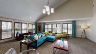 Located at 2020 Armstrong Mill Rd in Lexington, Kentucky, this single-floor clubhouse facility is designed to serve as a welcoming communal gathering space. The interior is defined by soaring vaulted ceilings that lend an airy, open feel throughout. A striking color palette of teal, yellow-green, and warm neutrals creates a lively yet cohesive aesthetic across all areas.

The lounge area is generously proportioned, anchored by a bold teal sectional sofa and complementary yellow accent chairs arranged around a central coffee table. A bank of large windows dressed with teal drape panels and horizontal blinds floods the space with natural light while offering views of the surrounding greenery. An elegant multi-pendant chandelier serves as a focal point overhead.

A dedicated media area features a teal accent wall with a wall-mounted flat-screen television and a rich wood media console, providing a comfortable secondary seating zone. Floor-to-ceiling glass partition walls connect this space to an adjacent dining and kitchen area, maintaining visual openness while defining distinct functional zones.

The dining area accommodates multiple seating arrangements with bar-height tables and mixed seating, flowing seamlessly into a full kitchen equipped with cabinetry and appliances. Textured, patterned carpet ties the various zones together, while recessed lighting and the central chandelier ensure well-illuminated interiors throughout.