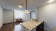 This single-floor unit offers a clean, modern layout with consistent dark hardwood-style flooring throughout. The open-concept kitchen serves as the centerpiece of the living area, anchored by a peninsula countertop finished in light granite with natural veining. Dark espresso cabinetry provides ample storage above, while black appliances — including a gas range, over-the-range microwave, and refrigerator — lend a sleek, uniform aesthetic. A pendant light with a classic cone shade hangs above the peninsula, adding a warm focal point to the space. The kitchen flows naturally into the living area, which benefits from natural light through a window fitted with horizontal blinds and features a wall-mounted HVAC unit. The bedroom is generously proportioned, offering a clean white canvas with a closet rod and window for natural light. The bathroom includes a wall-mounted toilet, a full-length framed mirror, built-in linen storage with a two-door cabinet, a towel bar, and a white countertop vanity. Throughout the unit, white walls and recessed lighting create a bright, neutral atmosphere that is move-in ready and easily personalized.