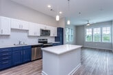 This single-floor residence in Charleston, South Carolina presents a thoughtfully designed interior with a cohesive modern aesthetic. The kitchen serves as a standout feature, showcasing a striking two-tone cabinet scheme pairing crisp white upper cabinets with bold navy lower cabinets, complemented by a subway tile backsplash and white quartz countertops. A central island provides additional prep and gathering space, while stainless steel appliances—including a refrigerator, range, microwave, and dishwasher—complete the functional layout. Pendant lighting overhead adds a polished finishing touch. The kitchen flows seamlessly into an open living area bathed in natural light from multiple windows fitted with white blinds. Light hardwood-style flooring extends throughout the main living spaces, enhancing the sense of openness, while a ceiling fan provides comfort year-round. The bedroom features plush carpeting and a generously sized window with views of surrounding greenery. The bathroom continues the navy cabinetry theme with a wide vanity topped in white, a round mirror, and a tub-shower combination with tiled surround, all set on wood-look flooring. Neutral gray walls throughout lend a calm, contemporary ambiance well-suited to Charleston living.