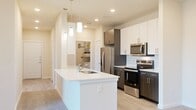 This well-appointed 3-bedroom, 3-bathroom single-story residence offers a modern and functional layout designed for comfortable living. The contemporary kitchen is a standout feature, showcasing a two-tone cabinetry scheme with white upper cabinets and dark wood-grain lower cabinets, sleek white quartz countertops, a white subway tile backsplash, and a full suite of stainless steel appliances. A peninsula with pendant lighting provides additional prep space and casual seating. Light wood-style flooring runs throughout the kitchen and main areas, while plush carpeting extends into the bedrooms and living spaces.

A carpeted hallway connects the living areas to the bedrooms, each equipped with ceiling fans and ample natural light from windows with blinds. The spacious primary bedroom features sliding glass doors that open to a private balcony with open views. Bedrooms include walk-in closets with built-in shelving for generous storage. The living room features built-in shelving and an arched accent detail, adding architectural character to the space.

A dedicated laundry area houses a full-size washer and dryer with an overhead water heater. Three full bathrooms and two closets round out this efficiently designed residence, offering a balance of style and practicality throughout.