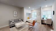This single-floor studio is designed to maximize every square foot through smart, built-in solutions and a cohesive modern aesthetic. The open-concept layout seamlessly connects the living, sleeping, and kitchen areas, all unified by light wood-tone flooring and a neutral gray-and-white color palette throughout.

The sleeping area is anchored by a built-in bed nook framed with floor-to-ceiling white cabinetry, offering generous overhead storage while keeping the space feeling organized and intentional. A window adjacent to the bed draws in natural light, brightening the nook. The living area accommodates a gray sectional sofa, a white coffee table, and a leather accent chair, creating a comfortable and stylish lounge space.

A dedicated workspace along one wall features a built-in desk and is complemented by additional open shelving, making it well-suited for remote work or study.

The kitchen is fully equipped with stainless steel appliances — including a full-size refrigerator, dishwasher, oven, and microwave — alongside white shaker cabinetry and a tile backsplash. An in-unit washer is conveniently integrated into the kitchen area.

The bathroom is finished with clean white subway tile in the shower-tub combo, gray tile flooring, and brushed nickel fixtures, delivering a polished, contemporary feel.