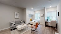 This single-floor studio is designed to maximize every square foot through smart, built-in solutions and a cohesive modern aesthetic. The open-concept layout seamlessly connects the living, sleeping, and kitchen areas, all unified by light wood-tone flooring and a neutral gray-and-white color palette throughout.

The sleeping area is anchored by a built-in bed nook framed with floor-to-ceiling white cabinetry, offering generous overhead storage while keeping the space feeling organized and intentional. A window adjacent to the bed draws in natural light, brightening the nook. The living area accommodates a gray sectional sofa, a white coffee table, and a leather accent chair, creating a comfortable and stylish lounge space.

A dedicated workspace along one wall features a built-in desk and is complemented by additional open shelving, making it well-suited for remote work or study.

The kitchen is fully equipped with stainless steel appliances — including a full-size refrigerator, dishwasher, oven, and microwave — alongside white shaker cabinetry and a tile backsplash. An in-unit washer is conveniently integrated into the kitchen area.

The bathroom is finished with clean white subway tile in the shower-tub combo, gray tile flooring, and brushed nickel fixtures, delivering a polished, contemporary feel.