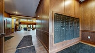 Located at 3090 Hazelwood St in Maplewood, Minnesota, this generously sized community common area is designed to impress with a warm, lodge-inspired aesthetic throughout. The space is anchored by a dramatic living room boasting soaring double-height ceilings, rich wood-paneled walls, and wood-look tile flooring that flows seamlessly through each area. A stunning floor-to-ceiling stacked stone fireplace serves as the living room's focal point, complemented by built-in shelving and wall sconces that add a refined ambiance. Large windows and glass doors flood the room with abundant natural light and offer inviting views of surrounding greenery, creating a bright and airy atmosphere. An adjoining kitchen area features warm wood cabinetry set against a stacked stone accent wall, complete with stainless steel appliances, a sink, and counter seating — ideal for gatherings and community events. The hallway serves as a functional and well-appointed corridor, finished with the same wood paneling and tile flooring, and equipped with individual mailboxes and a modern package locker system for resident convenience. Coffered ceiling details and carefully placed lighting throughout enhance the overall sense of quality and craftsmanship, making this a welcoming hub for the community.