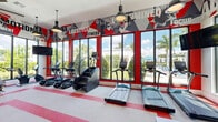 Located at Waterfront Crossing in Lake Worth, Florida, this fitness center delivers a comprehensive workout environment across a single, thoughtfully divided floor. The main exercise area is defined by its bold, high-energy aesthetic — vibrant red and gray motivational graphics and athletic murals cover the walls, creating an immersive, energizing atmosphere. Large floor-to-ceiling windows line the perimeter, flooding the space with natural light and offering refreshing views of tropical landscaping and palm trees beyond. The main floor is outfitted with a full suite of strength and cardio equipment, including cable machines, power racks, weight benches, treadmills, stair climbers, ellipticals, and stationary bikes, all arranged on durable, striped performance flooring. Industrial pendant lighting accents the space with a modern touch, while wall-mounted flat-screen TVs provide entertainment throughout. A separate studio room adjoins the main floor, featuring warm hardwood floors, full-length mirrored walls, recessed ceiling lighting, and a dedicated flat-screen display. This quieter space is equipped with spin bikes and open floor area, making it ideal for cycling classes or focused training sessions. Together, the two areas form a versatile, well-appointed fitness facility suited for a wide range of workout routines.