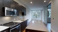 This contemporary single-floor space showcases a clean, modern aesthetic throughout its open-concept layout. The kitchen is a standout feature, designed with dark espresso cabinetry that reaches toward the ceiling, offering ample storage while creating a bold visual contrast against the crisp white quartz countertops. A subway tile backsplash in soft grey adds texture and depth to the cooking area, complementing the full suite of stainless steel appliances, including a built-in microwave, dishwasher, and range. A central kitchen island provides additional counter space and serves as a natural gathering point. The sink area is neatly integrated into the countertop run, keeping the workspace functional and streamlined. Warm hardwood flooring anchors the kitchen and flows seamlessly into the adjoining living area, where large picture windows bathe the space in abundant natural light and frame lush, green tree canopy views beyond. Recessed lighting overhead ensures the interior remains well-lit throughout. The overall ambiance is bright, polished, and inviting, with a thoughtful blend of dark and light tones that give the space a refined, contemporary character suited for modern living.