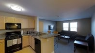 This single-floor space offers an open-concept layout that combines the kitchen and living area into one cohesive, functional environment. The kitchen is equipped with light wood cabinetry, a double-basin sink, granite-style countertops, and black appliances including a dishwasher and range. A peninsula with bar seating extends from the kitchen, providing an ideal spot for casual dining or entertaining while maintaining a clear sightline to the living area. The living space is carpeted and features a dark sofa, a low-profile coffee table, and a wall-mounted television, creating a practical and comfortable lounge area. Natural light enters through a pair of windows along the far wall, brightening the room and giving it an open, airy feel. The neutral gray walls and light-toned flooring in the kitchen contribute to a clean, modern aesthetic throughout. The thoughtful design of this space maximizes functionality while maintaining a welcoming atmosphere suitable for everyday living.