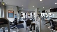 This fitness center is designed to support a well-rounded exercise routine in a clean, modern environment. The space is outfitted with a strong selection of cardio equipment, including multiple treadmills and elliptical machines, each equipped with individual display screens for entertainment and performance tracking. Strength training machines are also prominently featured, providing options for a variety of muscle groups and fitness levels. A wall of mirrors on one side of the room enhances the sense of openness while allowing users to monitor their form during workouts. Recessed ceiling lighting is evenly distributed throughout, ensuring the space remains bright and well-lit at all times. Large windows along one wall allow natural light to filter in and offer views of the surrounding exterior, contributing to an energizing atmosphere. The flooring includes both hard surfaces and rubberized mat areas, providing practical zones for different types of exercise. A water dispenser is conveniently positioned near the entrance, and informational signage is posted for facility guidelines. The overall ambiance is functional, modern, and well-maintained, making it a practical and inviting fitness facility.