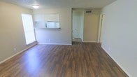 This single-floor space offers a practical and well-organized layout designed for comfortable everyday living. The main living area is anchored by rich, dark hardwood-style flooring that extends throughout, creating a warm and cohesive look underfoot. Natural light filters in through a window along the side wall, brightening the open space and highlighting the clean, neutral-toned walls.

A defining feature of the layout is the pass-through kitchen cutout, which connects the kitchen to the living area via a raised countertop and white cabinetry above. This design keeps the spaces visually linked while maintaining a clear functional separation. The kitchen is partially visible through the opening, featuring white appliances and a tiled or light-colored floor.

The textured ceiling adds a classic touch throughout the space, while a centrally located HVAC vent ensures climate comfort. Multiple doorways visible from the main living area lead to additional rooms, suggesting a well-connected and navigable floor plan. Baseboards and door trim are finished in crisp white, providing clean contrast against the warm walls. Overall, the space presents a tidy, move-in-ready feel with a straightforward, efficient design.