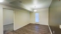 This single-floor space offers a clean, functional layout designed with simplicity and practicality in mind. The interior is finished with rich, dark hardwood-style flooring that runs throughout, providing a warm and grounded aesthetic that contrasts beautifully with the soft, neutral cream-toned walls. The textured ceiling adds a subtle architectural detail overhead, while crisp white baseboards frame the perimeter of each room, contributing to a polished, well-maintained appearance. A standout feature of the main living area is an elegant pendant light fixture that hangs centrally, casting warm ambient light across the open space. A door with a windowed panel allows natural light to filter in, brightening the interior and offering a visual connection to the outside. The layout provides a sense of openness, with clean sightlines and minimal obstructions that make the space feel airy and easy to navigate. Additional doorways lead to adjoining rooms, suggesting a well-organized and accessible floor plan. Overall, the space presents a neutral, move-in-ready environment that is versatile and well-suited for a variety of uses.