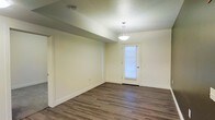This single-floor space offers a clean, functional layout designed with simplicity and practicality in mind. The interior is finished with rich, dark hardwood-style flooring that runs throughout, providing a warm and grounded aesthetic that contrasts beautifully with the soft, neutral cream-toned walls. The textured ceiling adds a subtle architectural detail overhead, while crisp white baseboards frame the perimeter of each room, contributing to a polished, well-maintained appearance. A standout feature of the main living area is an elegant pendant light fixture that hangs centrally, casting warm ambient light across the open space. A door with a windowed panel allows natural light to filter in, brightening the interior and offering a visual connection to the outside. The layout provides a sense of openness, with clean sightlines and minimal obstructions that make the space feel airy and easy to navigate. Additional doorways lead to adjoining rooms, suggesting a well-organized and accessible floor plan. Overall, the space presents a neutral, move-in-ready environment that is versatile and well-suited for a variety of uses.