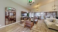 This two-story community room in Alpine, California, offers a well-appointed gathering space designed for socializing and recreation. The main floor showcases soaring vaulted ceilings that lend an open, airy atmosphere, with warm light wood flooring flowing throughout. The living area is furnished with comfortable lounge seating, a large flat-screen TV on a rustic media console, and arched accent mirrors that add visual depth. Elegant pendant chandeliers provide warm ambient lighting, while multiple windows dressed with wood-trimmed blinds and drape panels fill the space with natural light and views of the surrounding greenery. A dedicated dining nook features bistro-style seating and high-backed upholstered chairs, ideal for casual gatherings. The kitchen is equipped with rich dark wood cabinetry, granite countertops, a breakfast bar with wooden stools, and stainless steel appliances. A water station and restroom facilities are conveniently located nearby. The upper floor hosts the game room, carpeted for comfort and featuring a full-size pool table, shuffleboard table, a wall-mounted TV, pub-height seating, and ample natural light from multiple windows. Together, both floors create a versatile, inviting community space suited for a wide range of social activities.