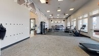 Located at 801 Watkins View Dr in Midlothian, Virginia, this well-appointed fitness center offers a comprehensive workout environment across a single open floor. The main exercise area features a bold geometric accent wall in gray and white tones, providing a striking visual backdrop to a wide array of strength and cardio equipment, including cable machines, rowing ergometers, free weights, and dumbbell racks. Ceiling fans and recessed lighting keep the space bright and comfortable throughout. A row of treadmills and cardio machines lines a bank of large windows and glass doors that open directly to a resort-style outdoor pool area, flooding the space with natural light and scenic outdoor views. An adjacent dedicated studio room offers wood-look flooring, large wall-mounted mirrors framed in dark trim, and patterned wallcovering, making it ideal for group fitness, cycling, and functional training. This studio also houses spinning bikes, a punching bag, a training zone rack, and stability balls. The overall design balances a clean, modern aesthetic with practical, high-capacity fitness functionality, creating an inviting and motivating environment for a wide range of workout routines.