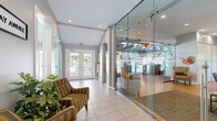 Located at 1010 N Knollwood Dr in Schaumburg, Illinois, Woodbury Place offers a thoughtfully designed single-story community facility that serves as a central hub for residents. The entrance features a bright, tile-floored lobby with floor-to-ceiling glass partitions that provide clear sightlines into the adjacent common areas, creating an open and transparent atmosphere. Comfortable seating and lush greenery near the entry establish a welcoming first impression.

The main gathering space is a standout, showcasing dramatic vaulted ceilings with exposed dark wood beams and trusses that lend a distinctive architectural character. A floor-to-ceiling natural stone fireplace serves as the room's focal point, complemented by hardwood-style flooring, a glass coffee table, and ample natural light streaming in through large sliding glass doors that open to outdoor views.

The fully equipped kitchen features warm wood cabinetry, dark granite countertops, a farmhouse-style sink, subway tile backsplash, and black appliances. A center island with bar seating makes the space highly functional for group gatherings. An adjacent dining area provides additional seating with a mix of tables and modern chairs.

A separate, more formal meeting or activity room offers recessed lighting, hardwood-style floors, and large windows, making it a versatile space suited for a variety of community programming and events.