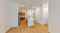 This thoughtfully designed one-bedroom, one-bathroom residence offers a cohesive blend of modern aesthetics and functional living across a single floor. The open-concept kitchen serves as a central feature, showcasing warm natural wood cabinetry, a classic subway tile backsplash, and stainless steel appliances including a refrigerator, range, and over-the-range microwave. A large white waterfall-edge island with a built-in sink anchors the space, illuminated by stylish pendant lighting overhead. Light hardwood flooring flows throughout the main living areas, lending warmth and continuity to the open layout. A stacked washer and dryer unit is conveniently integrated into the living area. The bedroom offers a comfortable, carpeted retreat with a ceiling fan and a window fitted with blinds that allows for natural light. The bathroom is a standout feature, presenting a double-sink vanity with crisp white countertops, natural wood cabinetry, matte black fixtures, and a large framed mirror with contemporary sconce lighting. Built-in shelving adds additional storage options. The walk-in closet is generously proportioned with wrap-around shelving and hanging rods, providing ample organization space. Overall, the space presents a clean, modern design with warm wood tones and bright white walls.
