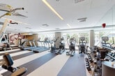 Located at 1440 Beacon St in Boston's Brookline neighborhood, this well-equipped fitness center offers a thoughtfully designed workout environment across a single floor. The space features a clean, modern aesthetic with a bright white ceiling fitted with recessed and linear LED lighting, complemented by large floor-to-ceiling windows along one wall that flood the room with natural light and provide views of the surrounding greenery.

The cardio zone is prominently featured and includes multiple treadmills, elliptical trainers, and stationary bikes, all positioned to take advantage of the natural light from the expansive windows. A dedicated free weights area offers a comprehensive rack of dumbbells in a range of sizes, providing ample options for strength training. Toward the rear of the space, a power rack and cable machine anchor the strength training section, set against a striking decorative wall mural featuring an abstract botanical pattern in deep burgundy and white tones, serving as a bold visual focal point.

The flooring transitions between light and dark striped fitness mats and rubberized workout zones, clearly delineating different training areas. Built-in shelving and storage add functional convenience. Full-length mirrors on one wall enhance the sense of space while supporting proper form during exercise. The overall ambiance is clean, professional, and energizing.