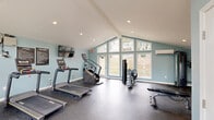 Located at 7303 Bryn Athyn Way in Raleigh, NC, this dedicated fitness center is thoughtfully designed to provide a bright, open, and motivating workout environment. The room's most striking architectural feature is its vaulted ceiling, which rises dramatically overhead and is complemented by a large, multi-panel window wall that spans nearly floor to ceiling. This expansive glazing floods the space with natural light and offers views of the surrounding greenery, creating an airy and energizing atmosphere.

The interior is finished with soft blue-green walls, white trim, and recessed lighting, lending the room a clean and modern aesthetic. Durable rubber-flecked flooring covers the entire space, providing cushioning and traction ideal for athletic use. A dedicated entry door with keypad access ensures secure, convenient entry for members.

The fitness center is well-equipped with multiple treadmills, a stair climber, a cable strength training machine, a multi-function weight machine, and a variety of free weights and accessories. A wall-mounted television is positioned for easy viewing during cardio sessions. The combination of quality equipment, soaring ceilings, and generous natural light makes this fitness center a functional and inviting space for residents to pursue their health and wellness goals.