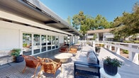 Located at 444 Saratoga Ave in Santa Clara, California, this single-story community space is designed to blend indoor comfort with expansive outdoor living. The centerpiece of the property is a generously sized open-air deck constructed with wide-plank composite decking, offering multiple distinct seating zones suited for both casual relaxation and social gatherings. A partially covered pergola structure with clean-lined columns provides shade over a portion of the deck, creating a transitional space between interior rooms and the outdoors. The covered area features wicker and rattan lounge chairs alongside a deep-seated sofa set, all arranged around low accent tables atop a patterned area rug. White cable-rail fencing borders the deck perimeter, adding a modern, open feel while framing views of the surrounding landscaped grounds. Mature trees envelop the space, lending a sense of privacy and natural shade under clear California skies. Additional dining-height tables and chairs are positioned throughout the deck for flexible use. Stone accent walls visible near the interior entry points add texture and warmth to the overall aesthetic. The floor plan reveals an L-shaped building footprint with interior spaces opening directly onto the deck through large sliding glass doors, ensuring seamless indoor-outdoor flow throughout.