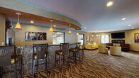 This single-floor hospitality space is thoughtfully designed to serve both social and functional needs. The layout is divided into distinct yet open zones that flow naturally into one another, unified by a bold, wave-patterned carpet throughout the main area.

The centerpiece of the space is a curved bar counter with a textured panel base, topped with a dark surface and surrounded by stylish high-back bar stools with green upholstered seats. Three amber pendant lights hang above the bar, adding warmth and visual interest. A curved architectural soffit overhead defines the bar zone and adds dimension to the ceiling design.

Adjacent to the bar, a comfortable lounge area features yellow upholstered sofas and accent chairs, arranged around a wall-mounted flat-screen television. Decorative artwork adorns the textured walls, and multiple windows allow natural light to fill the space. Additional seating options, including a brown leather chair and high-top table sets, offer flexible arrangements for guests.

The kitchen area is accessed behind the bar and features dark cabinetry with a tile backsplash, dark stone countertops, a stainless steel side-by-side refrigerator, a built-in microwave, a sink, and a coffee maker, making it fully equipped for hospitality service. Wood-look tile flooring grounds the kitchen zone distinctly from the carpeted lounge.