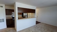 This single-floor residence showcases a practical and well-organized layout centered around an open-concept design. The kitchen is a standout feature, defined by a built-in pass-through counter with a light stone-toned surface that connects the cooking area to the adjacent living space. Dark espresso-toned upper cabinetry frames the pass-through opening, creating a bold visual contrast against the white walls. Within the kitchen, warm wood cabinetry provides ample storage, while stainless steel appliances — including a refrigerator and range — offer a modern, functional touch. The kitchen also features a dedicated sink positioned along the counter, enhancing its usability. The living area is finished with neutral carpeting that contributes to a calm, understated atmosphere, while the kitchen side transitions to dark hardwood-style flooring, clearly delineating the two zones. White walls and a clean, minimal aesthetic run throughout the space, lending it a bright and airy quality. A standard interior door is visible beyond the pass-through, suggesting additional rooms or access points within the residence. Overall, the space is designed for ease of movement and everyday functionality.