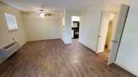 This single-floor residence offers a clean, straightforward layout designed for comfortable everyday living. The main living area is bright and open, finished with durable wood-look laminate flooring that runs throughout the space, creating a warm and cohesive feel. A ceiling fan is centrally mounted overhead, providing both light and air circulation. The neutral, light-toned walls keep the space feeling airy and versatile, ready to accommodate a variety of furnishing styles. A pass-through opening connects the living area to the kitchen, allowing for an open sightline between the two spaces. The kitchen is equipped with black appliances, including a range and overhead vent hood, complemented by white cabinetry that provides ample storage. The contrast between the dark appliances and light cabinetry gives the kitchen a classic, functional aesthetic. An interior door adjacent to the living area leads to additional rooms within the residence, suggesting a practical floor plan. The overall ambiance of the space is modest and functional, with clean finishes and a neutral palette that creates a blank canvas for personalization.