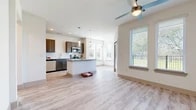 This single-floor unit presents a clean, contemporary design highlighted by light wood-style flooring and a crisp neutral color palette that runs consistently throughout the space. The open-concept layout connects the kitchen, dining, and living areas, creating a seamless flow ideal for everyday living and entertaining.

The kitchen is well-appointed with gray shaker-style cabinetry, a white subway tile backsplash, granite countertops, and a full suite of stainless steel appliances including a refrigerator, range, microwave, and dishwasher. A central island with a granite top provides additional workspace and casual seating. Pendant lighting above the island and recessed ceiling lights enhance functionality and ambiance.

The living area is generously proportioned, featuring a ceiling fan and multiple windows fitted with horizontal blinds that frame views of a landscaped green space and adjacent outdoor area, allowing abundant natural light. Additional rooms offer equally bright conditions, each with ceiling fans and large windows that bring in natural views.

The bathroom includes a tub-shower combination with large-format tile surround, a granite-topped vanity with gray cabinetry, and wood-style flooring that maintains the cohesive aesthetic found throughout the unit.