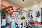 This contemporary conference room is a compact yet thoughtfully designed meeting space that blends functionality with striking artistic flair. The room's focal point is a bold, large-scale red abstract wall sculpture that spans the textured accent wall, lending an energetic and creative ambiance to the space. A wall-mounted flat-screen TV is integrated into this feature wall, making the room well-suited for presentations and collaborative work sessions.

At the center of the room, a light wood rectangular conference table is surrounded by modern ergonomic mesh chairs, comfortably seating up to eight people. A sculptural curved pendant light hangs above the table, providing focused illumination and serving as an additional design statement.

The flooring consists of patterned carpet tiles in muted tones accented with vibrant red insets that complement the room's color palette. Tall, sheer curtains frame large floor-to-ceiling windows, allowing abundant natural light to filter in while offering views of the surrounding exterior. Lush potted tropical plants are thoughtfully placed throughout, adding warmth and a touch of nature.

One side of the room features sleek floor-to-ceiling glass panel walls with black metal framing, offering visual connectivity to the adjacent lounge or reception area. This transparent partition enhances the sense of openness while maintaining a defined, private meeting environment.