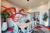 This contemporary conference room is a compact yet thoughtfully designed meeting space that blends functionality with striking artistic flair. The room's focal point is a bold, large-scale red abstract wall sculpture that spans the textured accent wall, lending an energetic and creative ambiance to the space. A wall-mounted flat-screen TV is integrated into this feature wall, making the room well-suited for presentations and collaborative work sessions.

At the center of the room, a light wood rectangular conference table is surrounded by modern ergonomic mesh chairs, comfortably seating up to eight people. A sculptural curved pendant light hangs above the table, providing focused illumination and serving as an additional design statement.

The flooring consists of patterned carpet tiles in muted tones accented with vibrant red insets that complement the room's color palette. Tall, sheer curtains frame large floor-to-ceiling windows, allowing abundant natural light to filter in while offering views of the surrounding exterior. Lush potted tropical plants are thoughtfully placed throughout, adding warmth and a touch of nature.

One side of the room features sleek floor-to-ceiling glass panel walls with black metal framing, offering visual connectivity to the adjacent lounge or reception area. This transparent partition enhances the sense of openness while maintaining a defined, private meeting environment.
