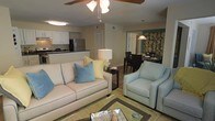This thoughtfully designed single-floor residence offers an open-concept layout that effortlessly connects the main living areas, creating a spacious and welcoming atmosphere throughout. The living room serves as the heart of the home, featuring a comfortable cream sofa accented with soft blue and yellow throw pillows, a plush light blue armchair, and a glass-topped coffee table. A ceiling fan overhead adds both function and a relaxed, casual charm to the space. Hardwood-style flooring extends throughout, lending warmth and visual continuity between rooms. The adjacent kitchen is well-appointed with white cabinetry, dark countertops, and stainless steel appliances, including a refrigerator and range, making it both practical and stylish. A bright overhead light keeps the workspace well-lit. Just beyond the living area, the dining space accommodates a dark wood table and chairs beneath a pendant light, creating an inviting setting for meals. A glimpse into an adjoining room reveals bold, patterned wallcovering, adding a touch of personality to the overall design. The open flow between spaces, consistent finishes, and cohesive coastal color palette give this residence a bright, airy, and livable quality throughout.