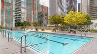 Located at 345 E Wacker Drive in the heart of Chicago, this outdoor amenity deck offers a compelling urban retreat set against a dramatic skyline backdrop. The centerpiece of the space is a generously sized lap pool with clearly marked lanes, framed by wide stone tile surrounds and stainless steel entry rails — ideal for both recreational and fitness-oriented swimming. Surrounding the pool, open terraces provide ample space for lounging and socializing, furnished with modern seating arrangements including chairs, sofas, and dining-height tables. Raised planters throughout the deck are filled with lush greenery, ornamental grasses, and colorful seasonal plantings, adding a natural contrast to the urban environment. A fully equipped outdoor kitchen station features a built-in stainless steel grill with a warm wood-paneled surround, making the space well-suited for outdoor entertaining. Floor-to-ceiling glass along the building's façade brings visual continuity between the indoor and outdoor areas, while the open terrace affords panoramic views of Chicago's iconic high-rise towers. The large-format stone tile flooring extends seamlessly across the entire deck, lending a cohesive, polished aesthetic to the entire outdoor amenity level.